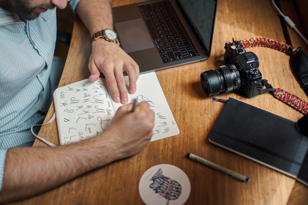 A man designing logos on a notebook next to a laptop and a camera.